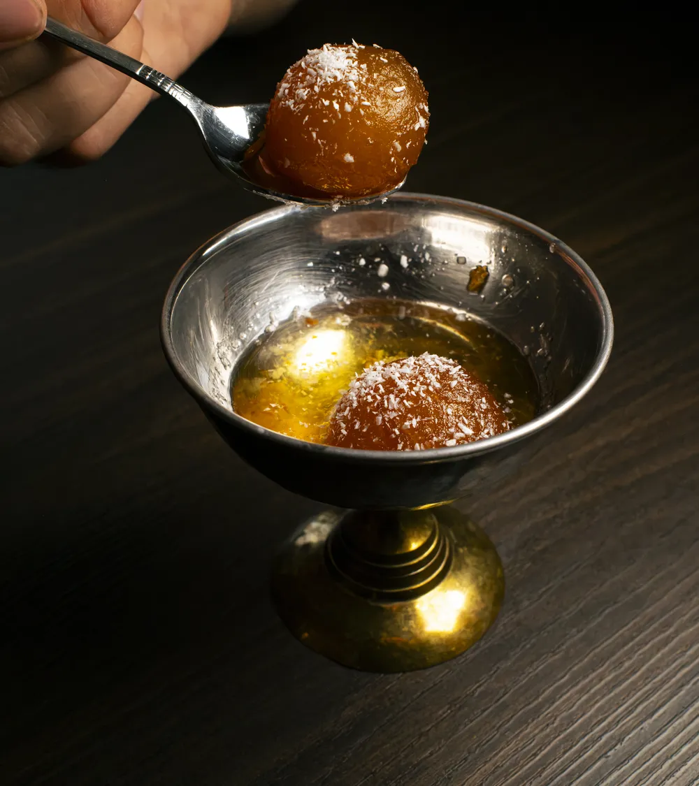 Un Gulab Jamun sollevato col cucchiaino dalla coppa in metallo